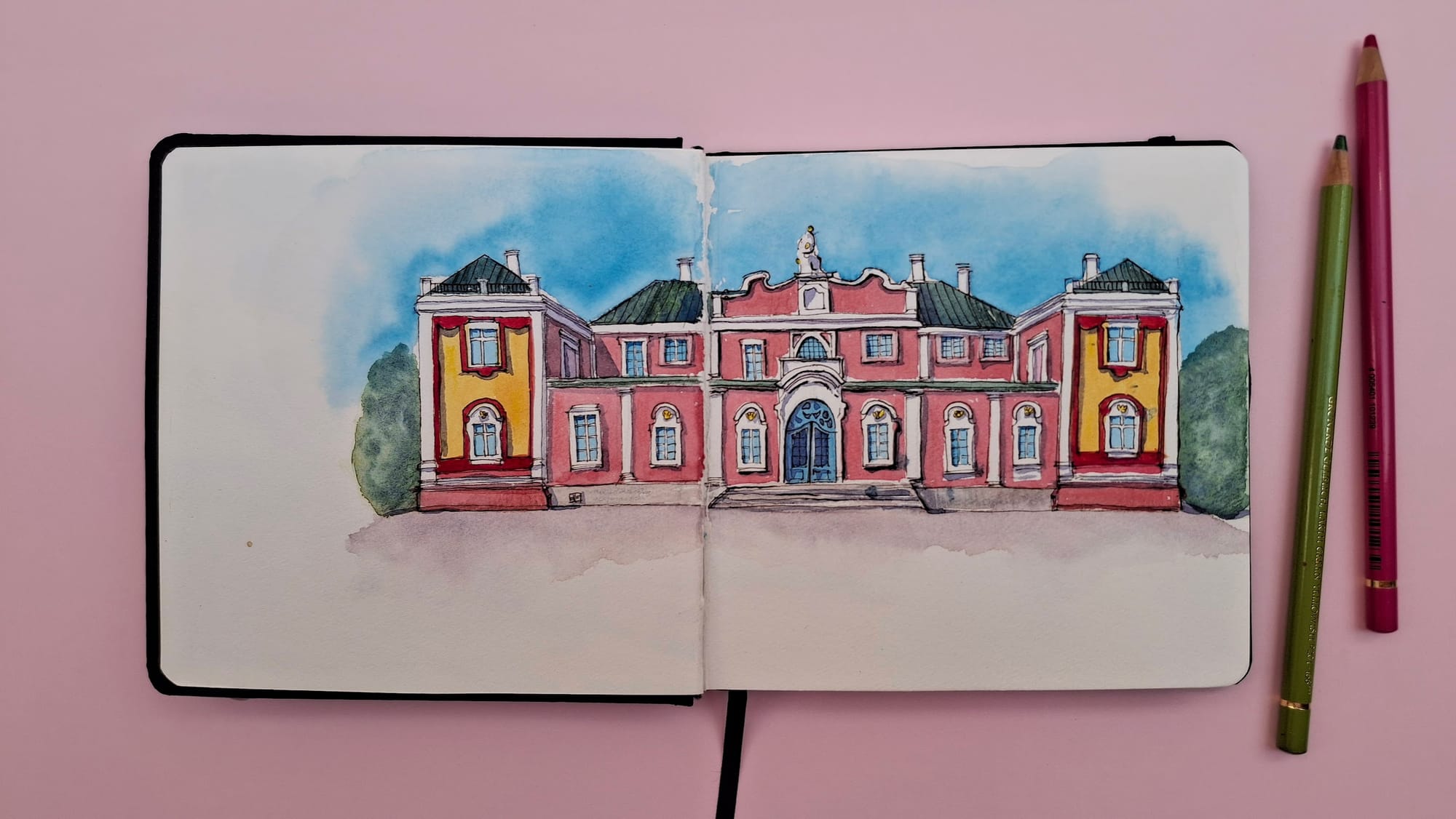A watercolour sketch of the Kadriog Art Museum (Kadrioru kunstimuuseum) in Tallinn, Estonia, by Emma Bolton. It's a two-storey pink building with green and yellow details, and is surrounded by greenery and a blue sky. The sketchbook is on a pale pink background with two coloured pencils. .