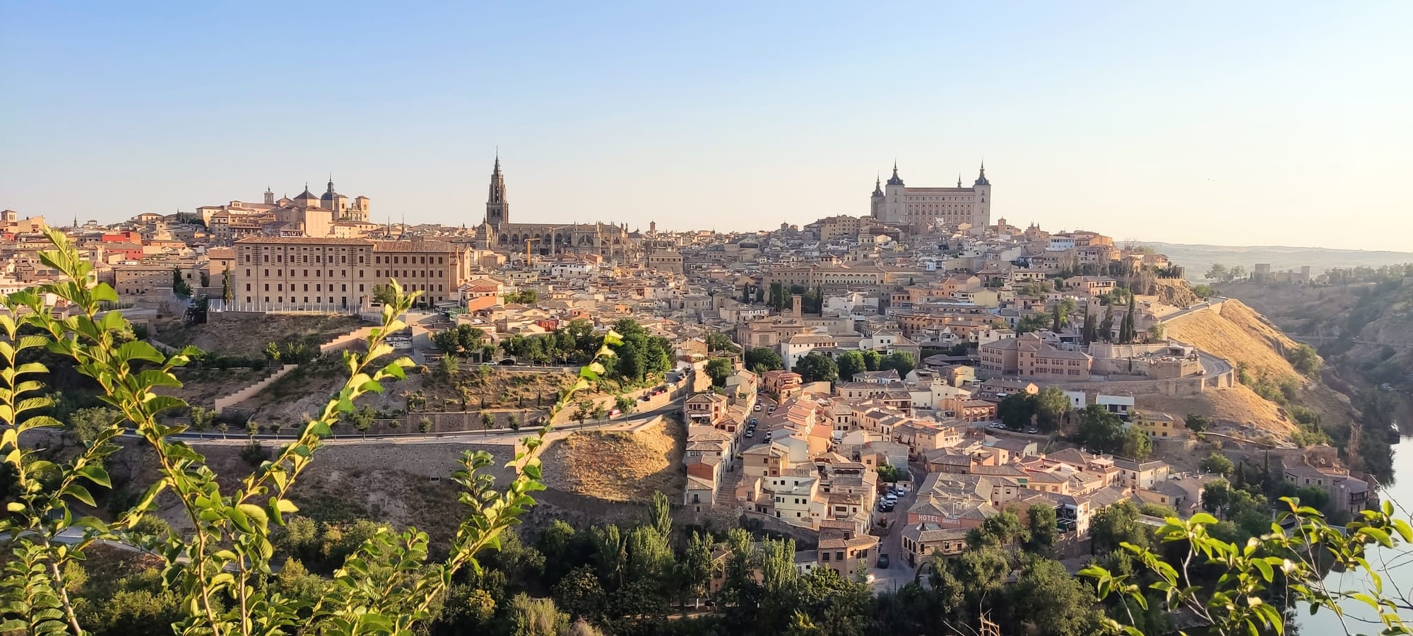 A view of Toledo, Spain from the top of an opposing hill. Sandstone buildings and greenery wind up the hill. A river is partially visible in the right corner.