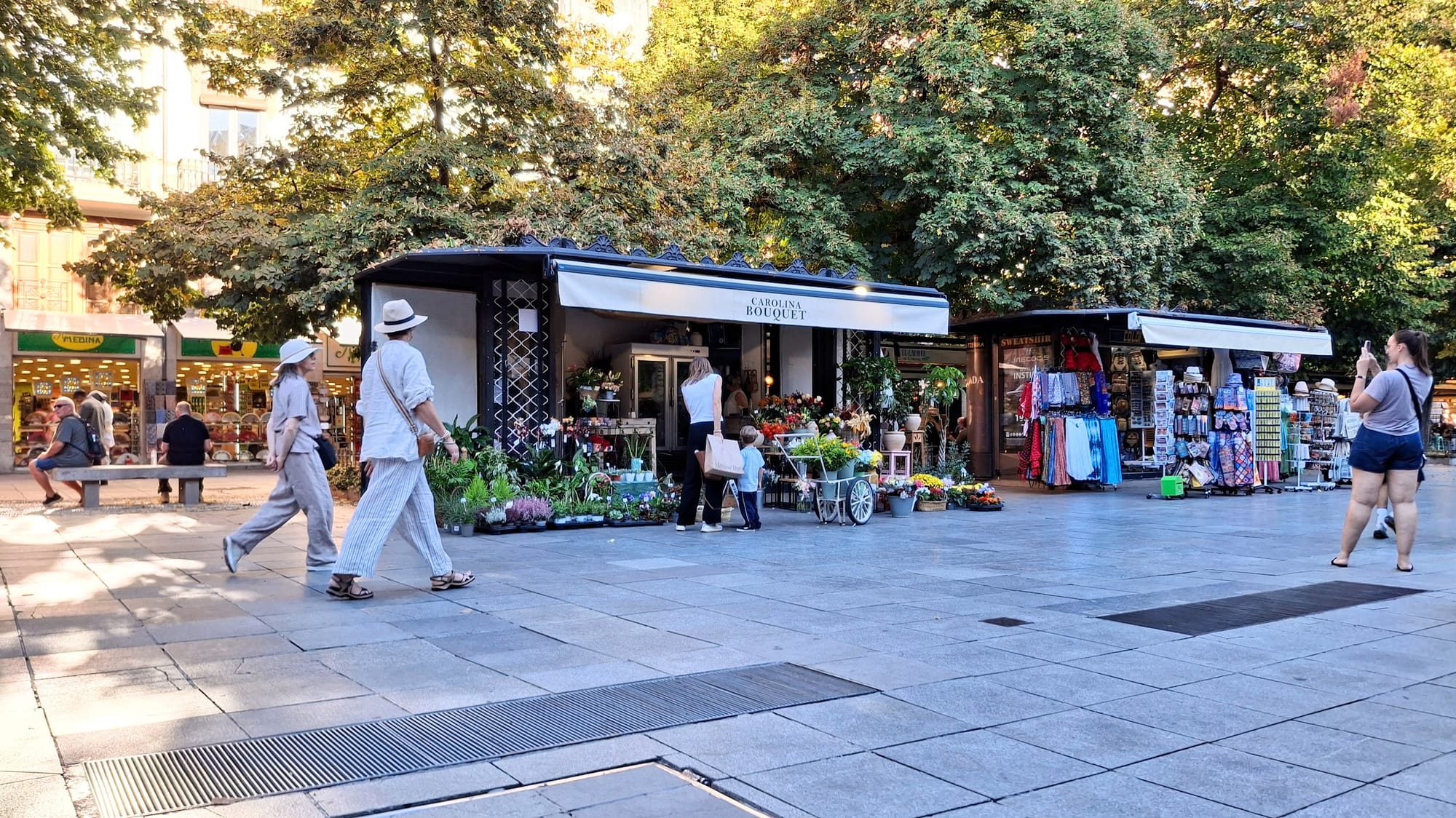 Carolina Bouquet in Bib Rambla, Granada. Various tourists are walking by and taking photos, and a woman and son are perusing the flowers. 