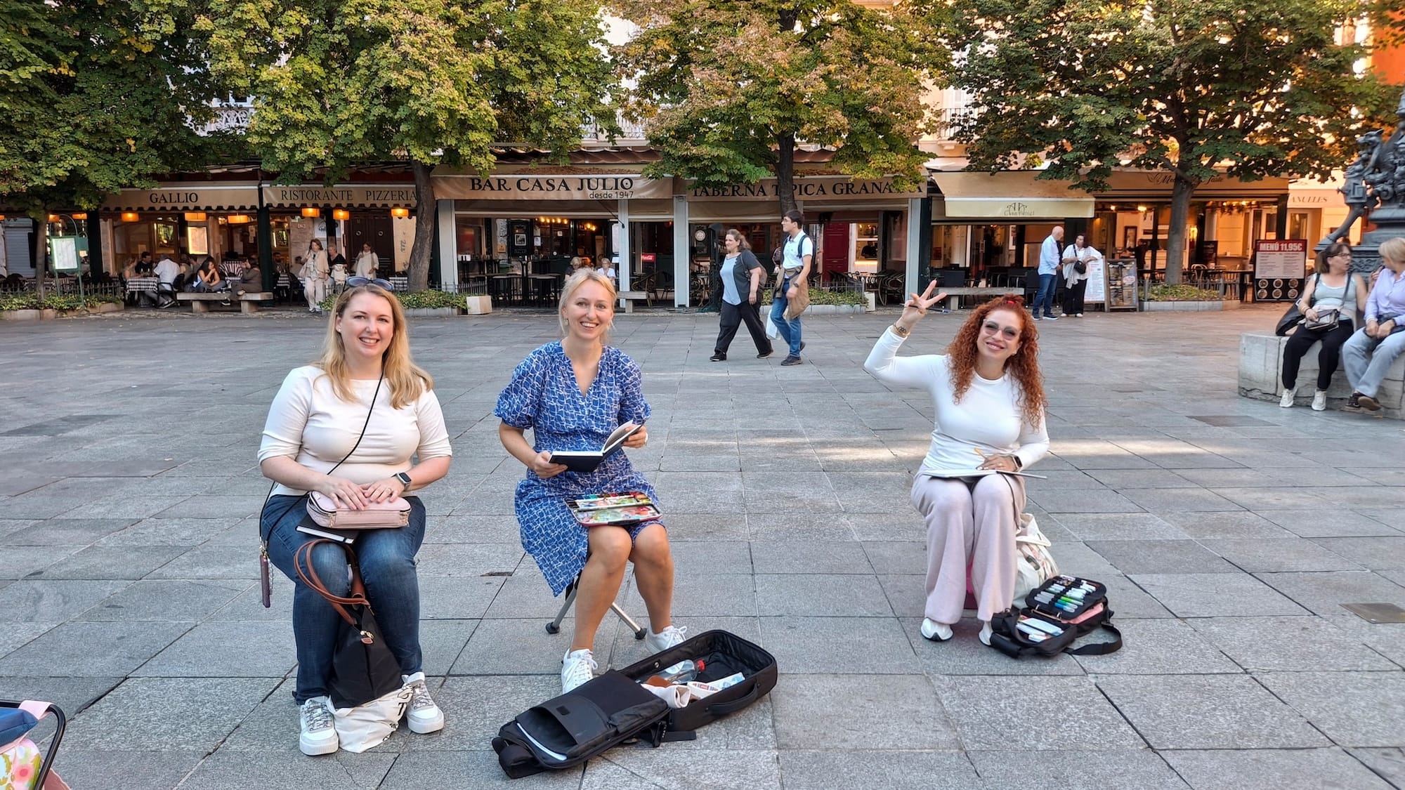 Three white women smiling and sitting on stools with their sketchbooks in the middle of Bib Rambla, Granada. In the background are restaurants, trees and a few tourists.