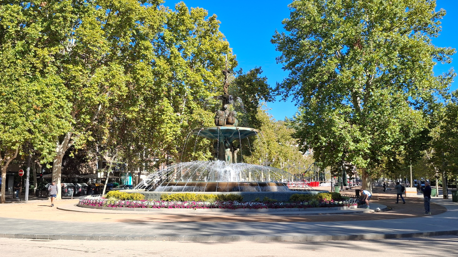 The three-tiered pomegranate fountain in central Granada with the water flowing in domes. Around are people walking and a leafy boulevard. 