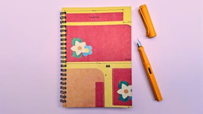 An A5 spiral notebook with red and yellow blocks and beige and pale blue flowers, together with a sunflower yellow Lamy fountain pen on a pale pink background.