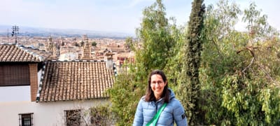 Emma Bolton wearing glasses, a blue puffer jacket and a green bag and smiling at the camera in the Albaicín.