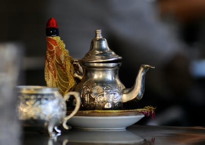 A silver engraved teapot with a gold and red cozy on the handle sits on top of a gold cushion and white plate. 