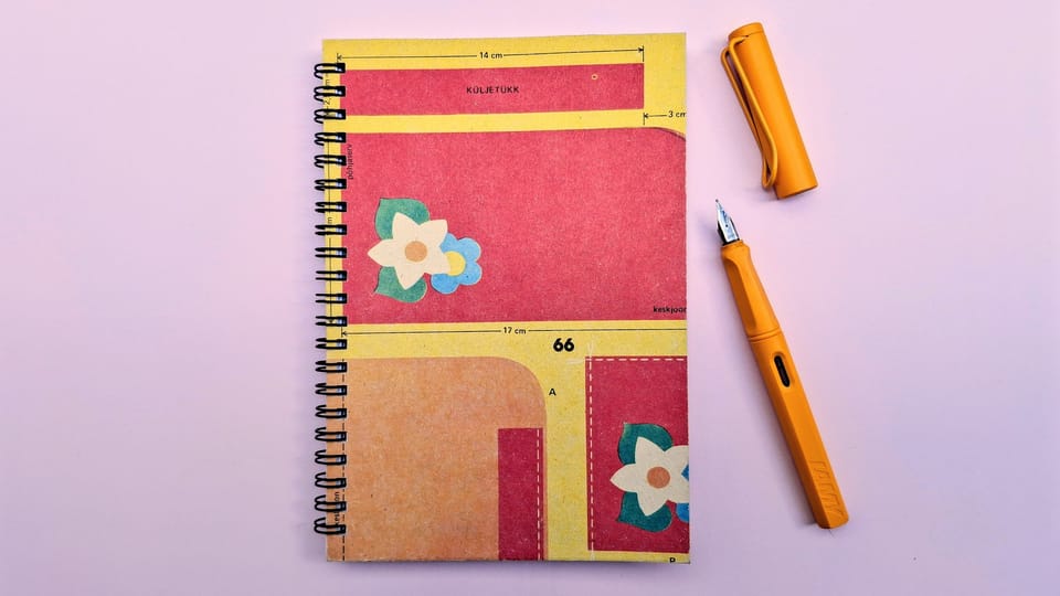 An A5 spiral notebook with red and yellow blocks and beige and pale blue flowers, together with a sunflower yellow Lamy fountain pen on a pale pink background.