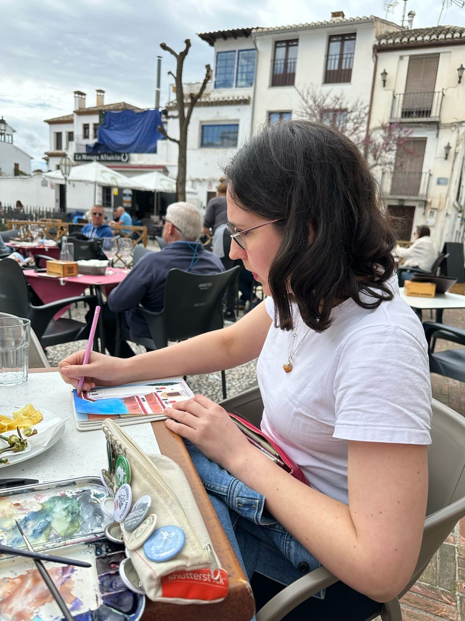 A white woman wearing glasses and a white t-shirt, sketching with a pink colored pencil at a café in Granada.