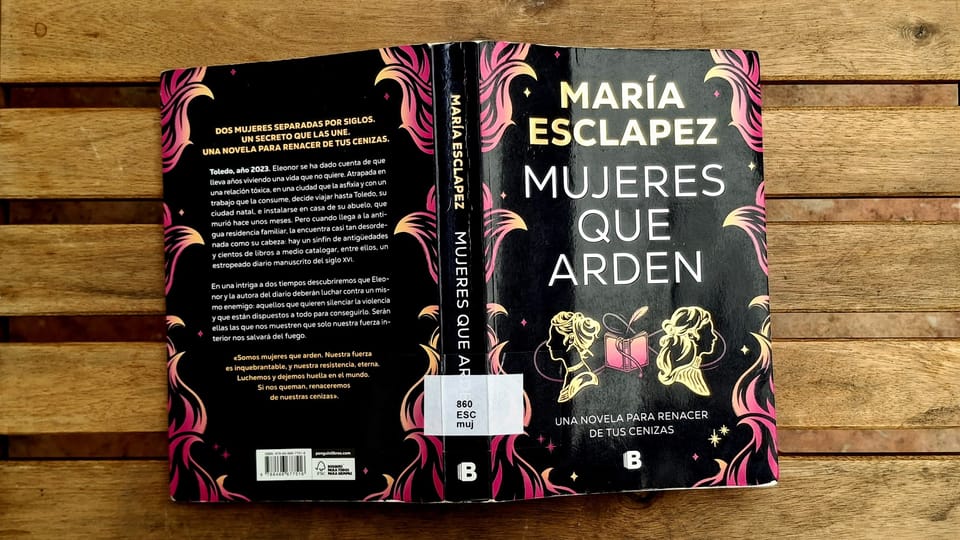 A copy of the book Mujeres que arden by María Esclapez on a wooden background. The book has a black cover with gold and pink details.