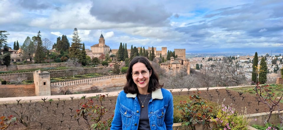 A white woman wearing glasses and a denim jacket is smiling at the camera. In the distance is the Alhambra palace and Granada.