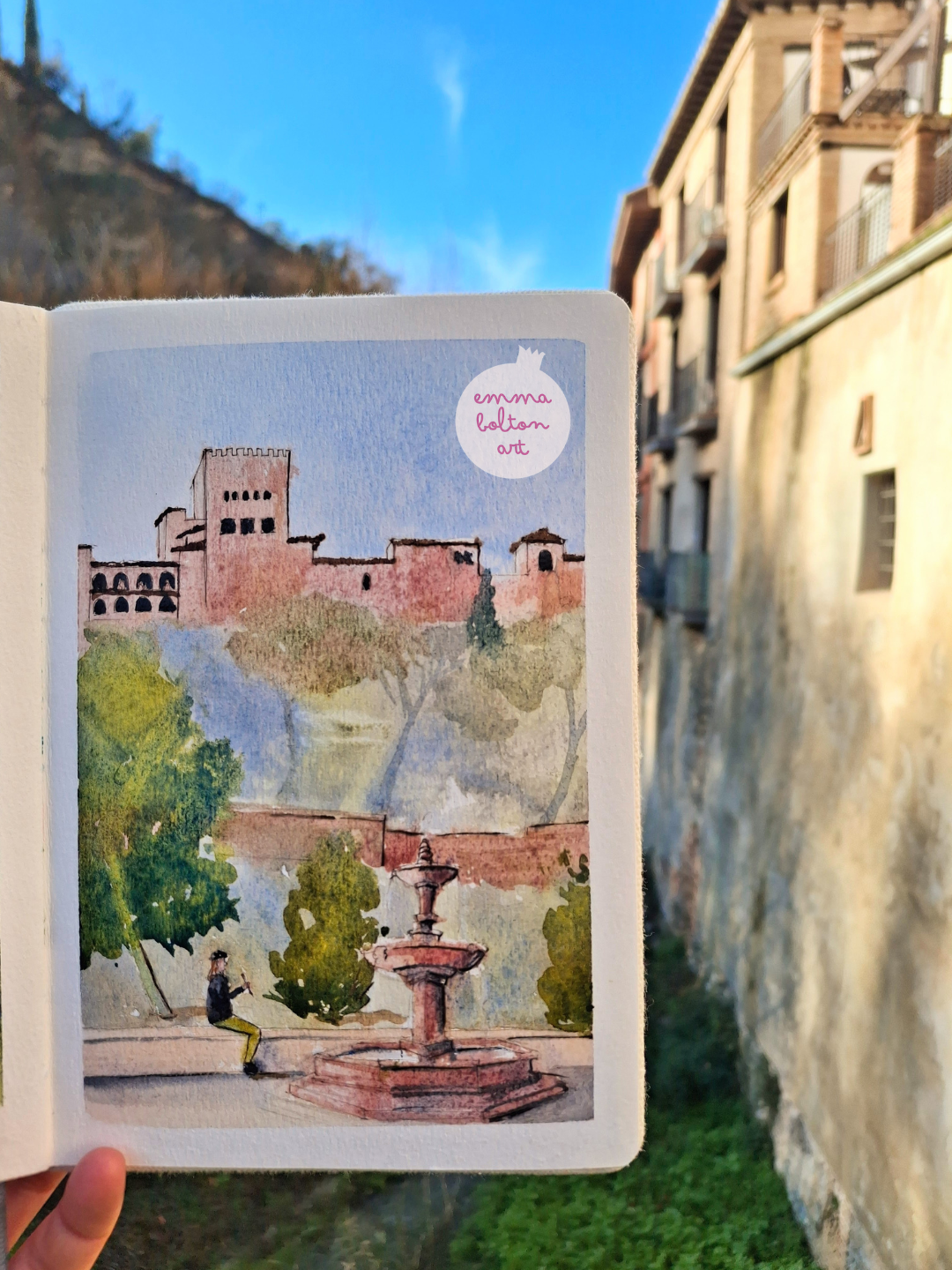 Watercolour sketch by Emma Bolton of the Alhambra, Granada and the fountain in Paseo de los Tristes. A man is sketching on a low wall. 