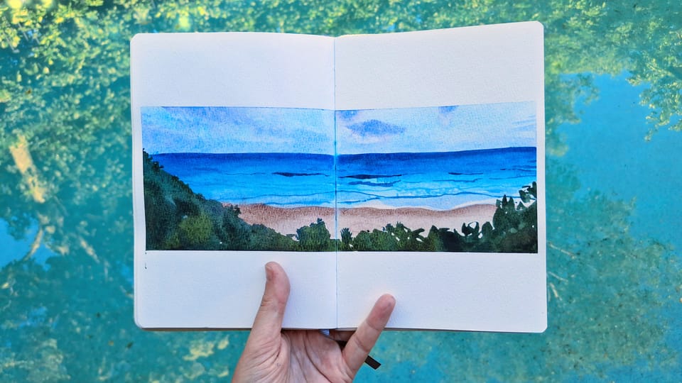 A hand holding an A5 sketchbook open over a pool with reflections of trees. The landscape sketch by Emma Bolton is of a turquoise ocean, pink sand and greenery in the foreground. 