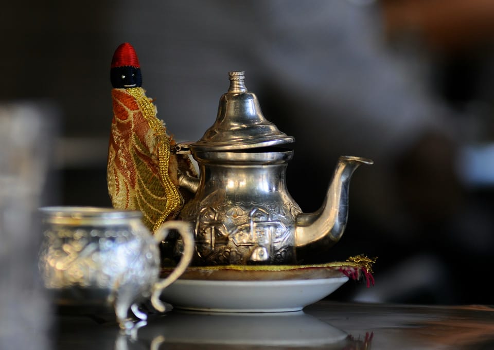 A silver engraved teapot with a gold and red cozy on the handle sits on top of a gold cushion and white plate. 